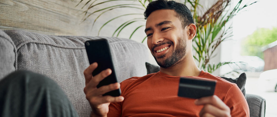 A man laying on a couch using a smartphone and holding a credit card